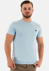 Ljusblå t-shirt med korta ärmar i bomull, med rund halsringning och en liten svart logga på bröstet. Diskret sidodetalj.