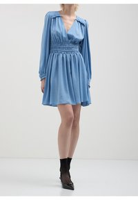 Robe bleu clair à manches longues, décolleté en V profond, taille smockée et jupe évasée. Présente une texture douce et une coupe décontractée. Chaussures noires à talons pointus.