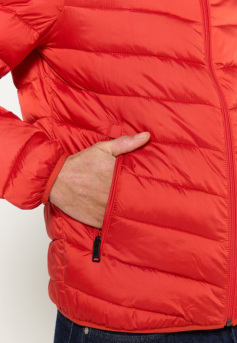 Chaqueta acolchada roja con costuras verticales, que presenta una mano en un bolsillo lateral, cierre de cremallera y puños elásticos. Textura suave y brillante.