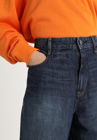 Granatowe dżinsy o wysokim stanem, z zapięciem na guzik i zamek błyskawiczny, z szlufkami na pasek i kieszeniami z przodu. W połączeniu z pomarańczowym swetrem.