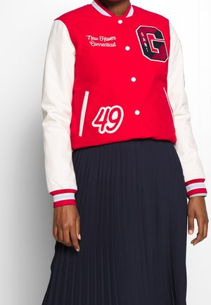 Rood-wit varsityjack met de teksten "New Haven Connecticut", "49" en een grote "G" patch, gedragen over een zwarte geplooide rok.
