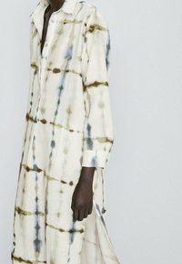 Vaaleanbeige kauluspaita-mekko, jossa on vihreä ja sininen batikkikuosi, pitkät hihat, joissa on napit ja sivureiät.