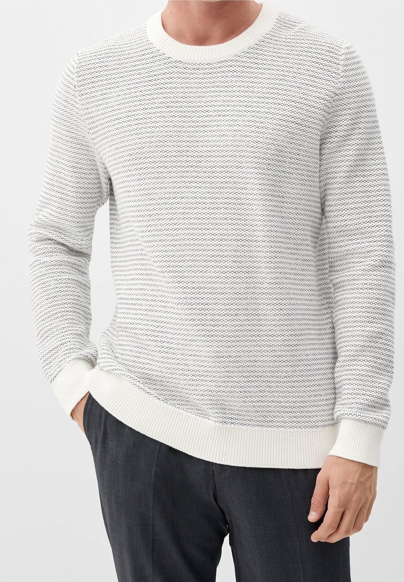 Homme portant un pull en maille texturée blanc et gris avec poignets et bas côtelés, associé à un pantalon gris foncé, main dans la poche.