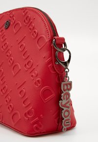 Sac synthétique texturé rouge avec un motif de logo embossé, fermeture éclair noire et un pendentif en métal portant l'inscription "Beyou."