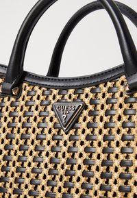 Sac à main tissé beige avec des accents et des poignées en cuir noir, arborant un logo noir en forme de triangle. Conçu avec un motif texturé et à mailles ouvertes.