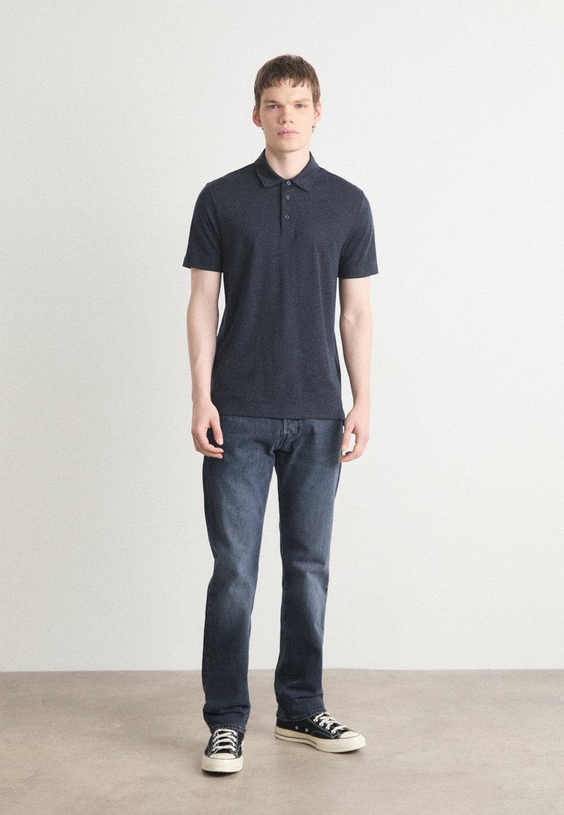 Marineblaues Poloshirt mit kurzen Ärmeln und einer Knopfleiste mit drei Tasten, kombiniert mit dunklen Jeans und schwarzen Turnschuhen. Schlichtes Design, lässige Passform.