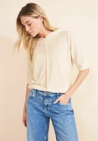 Beżowy sweter z dzianiny z kapturem i krótkimi rękawami, w połączeniu z jasnoniebieskimi dżinsami o klasycznym pięciokieszeniowym fasonie.
