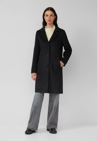 Schwarzer Wollmischmantel mit Reverskragen, drei Knöpfen und seitlichen Taschen, kombiniert mit hellgrauen, weit geschnittenen Jeans und schwarzen Schuhen.