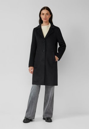 Schwarzer Wollmischmantel mit Reverskragen, drei Knöpfen und seitlichen Taschen, kombiniert mit hellgrauen, weit geschnittenen Jeans und schwarzen Schuhen.
