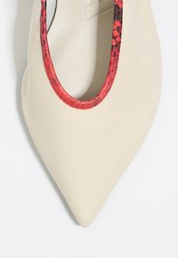 Zapato de cuero beige con puntera afilada y un ribete texturizado en rojo y negro alrededor de la abertura. Superficie suave con un ligero brillo.