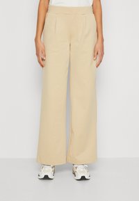 Beige wide-leg trousers with a smooth texture, featuring front pleats and an elastic waistband, paired with white and beige sneakers.