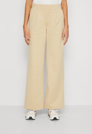 Beige vide bukser med en glat tekstur, der har frontfolders og en elastisk talje, parret med hvide og beige sneakers.