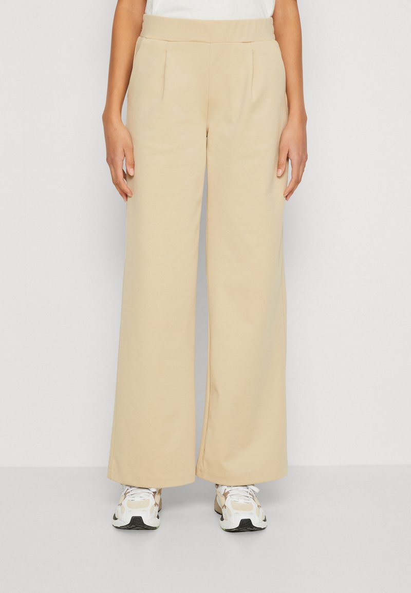 Beige leveled-hose dengan tekstur halus, dilengkapi dengan lipatan depan dan pinggang elastis, dipadukan dengan sepatu sneaker putih dan beige.