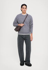 Grijze wollen trui, zwarte jeans met patchdetails en zwarte instapschoenen met bloemenborduurwerk. Bruine schoudertas. Neutrale achtergrond.