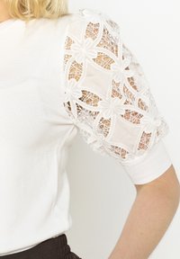 Vit stickad topp med spetspuffärmar med blommönster. Ärmarna har en ömtålig, strukturerad design och ribbade muddar.