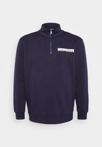 Granatowa bluza z zamkiem błyskawicznym i wysokim kołnierzem, z białym logo po lewej stronie klatki piersiowej oraz ściągaczami przy mankietach i dolnym brzegu. Miękka tekstura materiału.