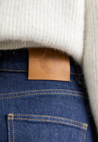 Tan leather tag with embossed floral pattern attached to denim jeans, paired with a light gray, ribbed knit sweater.