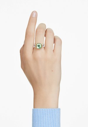 Mano con un anillo de oro con una gran piedra preciosa verde y pequeñas piedras laterales, llevado en el dedo medio sobre un puño azul claro.