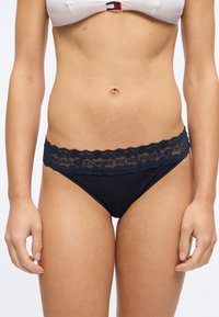 Γυναίκα που φορά εσώρουχα σε σκούρο μπλε με δαντέλα και λευκό σουτιέν, εμφανίζοντας τον κορμό και τα χέρια της μπροστά σε ένα απλό φόντο.