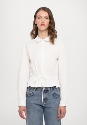 Weißes Hemd mit Knopfleiste und Kordelzug in der Taille; verfügt über lange Ärmel, einen Kragen und einen leicht strukturierten Stoff. Kombiniert mit blauen Jeans.