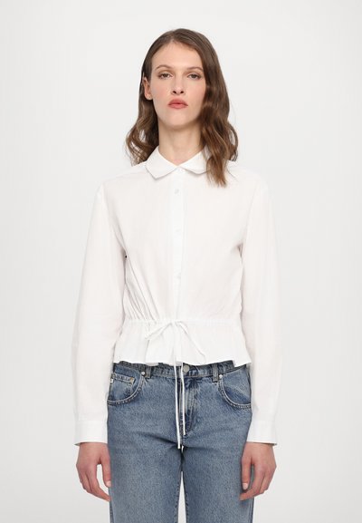 Chemise blanche à col boutonné avec ceinture à cordon ; elle présente de longues manches, un col et un tissu légèrement texturé. Associée à un jean bleu.