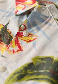 Camisa de manga corta de algodón con un patrón floral. Los colores dominantes son rojo, amarillo y verde, con un botón blanco en el cuello.
