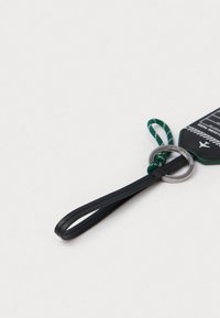 Fekete bőr kulcstartó, amely egy fémgyűrűhöz és egy zöld hurokhoz van rögzítve, egy fekete poggyász címkéhez csatlakozik, fehér felirattal.