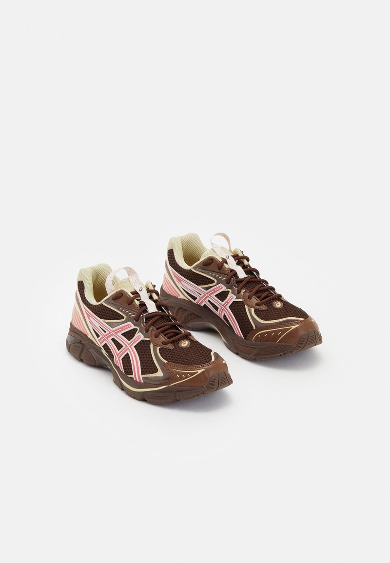 Ett par bruna och beige mesh löparskor med rosa och krämfärgade accenter och snören på en vit bakgrund.