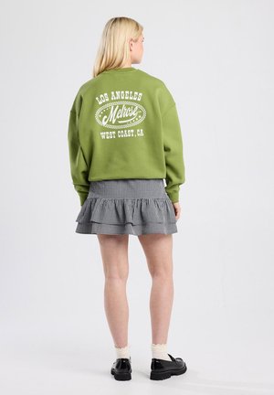 Groene sweatshirt met witte grafische tekst op de rug, gecombineerd met een zwart-wit geruite rok met franje. Zwarte schoenen en witte sokken maken de look compleet.
