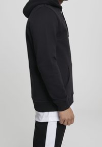 Svart hoodie med känguruficka, ribbade mudd och nederkant. Har en justerbar huva, visas från sidan, tillsammans med svarta byxor och en vit skjorta.