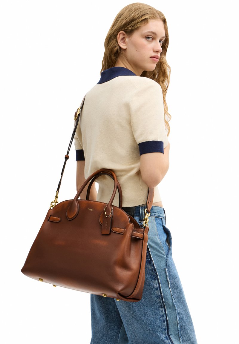 Jeune femme en haut beige et jean bleu portant un grand sac à main en cuir marron sur l'épaule, regardant par-dessus son épaule.