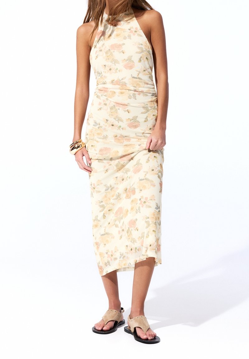 Femme portant une robe midi sans manches à fleurs avec des sandales beiges et des bracelets dorés au poignet gauche, debout sur un fond blanc.