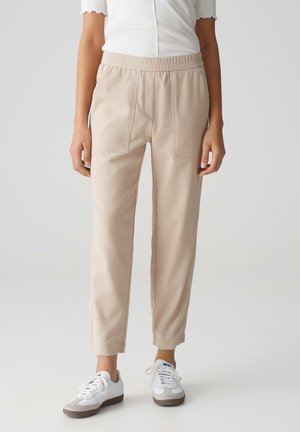 Beige, schmal zulaufende Hose mit elastischem Bund, zwei vorderen Taschen und glatter Textur, kombiniert mit weißen Sneakern.