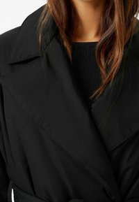 Svart trenchcoat med breda kraagar, tillverkad av slät tyg. Har en midja med bälte och en minimalistisk design som framhäver rena linjer och textur.