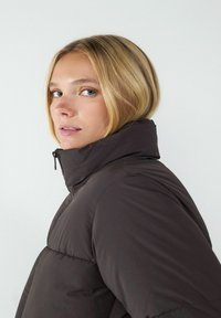 Dunkelbraune gepolsterte Jacke mit hohem Kragen und Reißverschluss. Glattes, wasserdichtes Material mit gesteppte Abschnitten zur Isolierung.