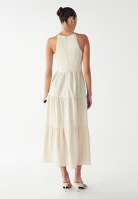 Nainen, jolla on nuttura ja yllään olkaimettomasta beigestä kerroksellisesta midi-mekosta sekä beigistä korkokengistä, seisoo yksinkertaista valkoista taustaa vasten.