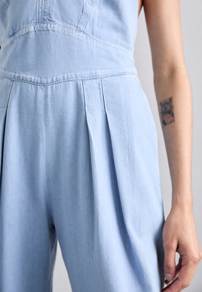 Salopette en denim bleu clair mettant en avant des plis à la taille, un corsage ajusté et un tissu texturé ; des détails de couture visibles rehaussent le design.