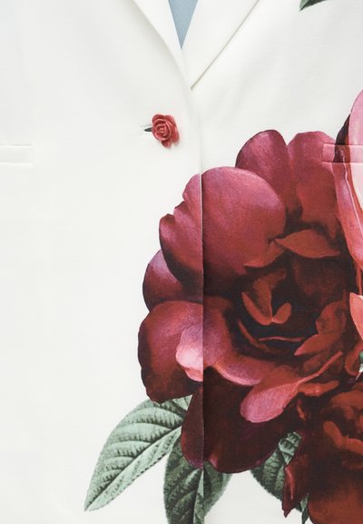Λευκό ένδυμα με μεγάλο λεπτομερές τύπωμα σκούρου κόκκινου λουλουδιού και κόκκινο κουμπί σε σχήμα τριαντάφυλλου κοντά στον γιακά.