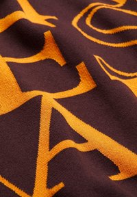 Stickad tyg i mörkbrun med stora, djärva orangea bokstäver. Slät textur, kantiga former och kontrasterande färger skapar en grafisk design.