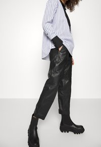 Gestreiftes blaues und weißes Oversize-Hemd mit schwarzen Manschetten; schwarze Lederhosen mit Nahtdetails; klobige schwarze Stiefeletten mit strukturiertem Sohlen.