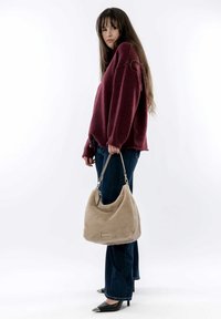 Sac hobo en fausse fourrure beige avec une texture lisse et des accessoires en argent, porté par une personne vêtue d'un pull bordeaux et d'un jean bootcut bleu foncé.