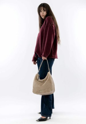 Sac hobo en fausse fourrure beige avec une texture lisse et des accessoires en argent, porté par une personne vêtue d'un pull bordeaux et d'un jean bootcut bleu foncé.