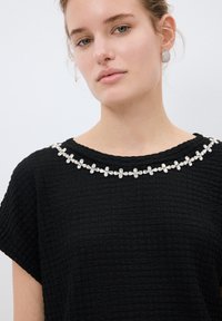Top nero in tessuto testurizzato con maniche corte, caratterizzato da un design del colletto floreale realizzato con gemme trasparenti incastonate in hardware argentato.