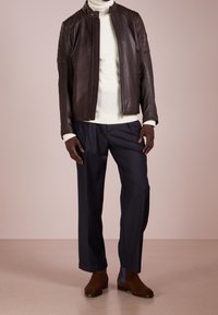 Brun läderjacka med vadderade axelpartier, bärs över en krämfärgad polotröja, ihop med marinblå byxor och bruna ankelstövlar.