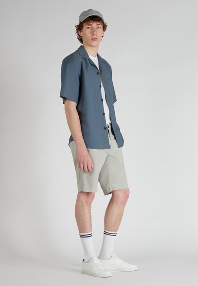 Jonge man staat, draagt een blauwe korte mouwen knoopshirt, lichtgrijze shorts, witte sokken met zwarte strepen, witte sneakers en een lichtgrijze pet.