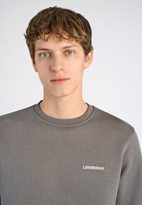 Sudadera gris con cuello redondo, que presenta un pequeño logotipo blanco "LINDBERGH" en el lado izquierdo del pecho. Textura suave y diseño minimalista.