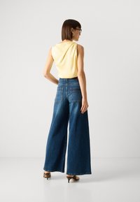 Top amarelo sem mangas combinado com calças de ganga de cintura alta e perna larga. As calças possuem dois bolsos traseiros e estão estilizadas com sapatos de salto castanhos.