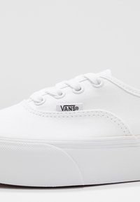 Białe płócienne trampki z okrągłym noskiem, metalowymi oczkami, białymi sznurowadłami i teksturowaną gumową podeszwą, z małym czarnym logo "VANS".