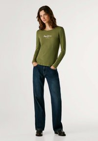 Langärmliges olivgrünes Hemd mit rundem Halsausschnitt und weißem "Pogo Jeans"-Logo, kombiniert mit dunkelblauen weit geschnittenen Jeans.