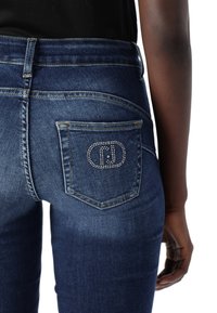 Vaqueros de mezclilla en azul oscuro con una vista en primer plano del bolsillo trasero, que presenta un logo engastado con diamantes y una tela texturizada.